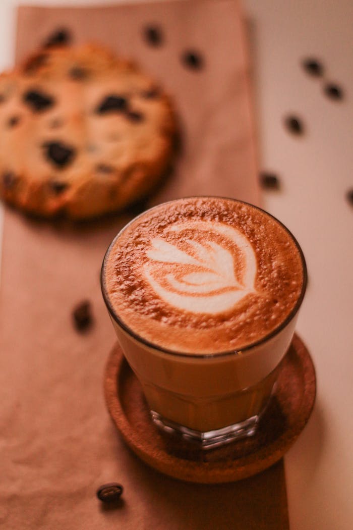journey A delicious latte with art foam beside a chocolate chip cookie on a wooden table, perfect for coffee lovers.