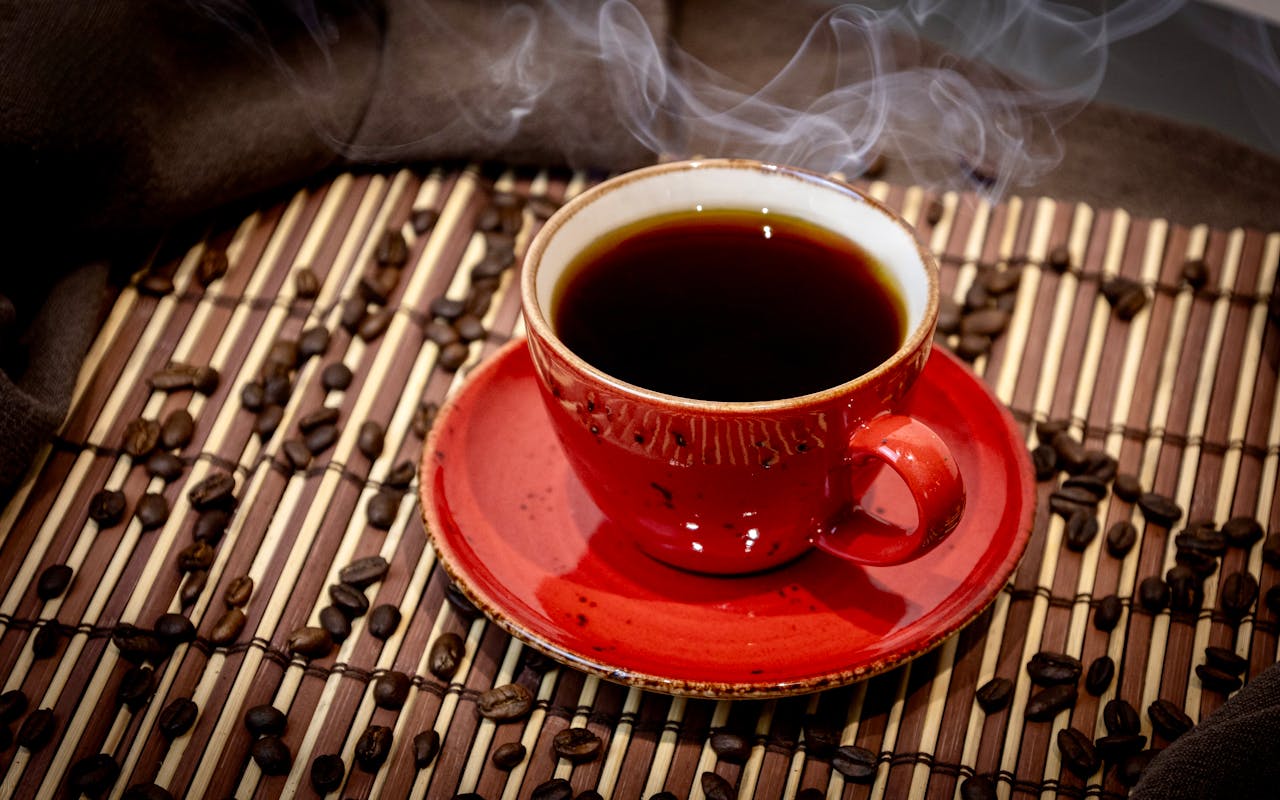 services-01 A steaming cup of coffee in a red mug surrounded by coffee beans on a bamboo mat.