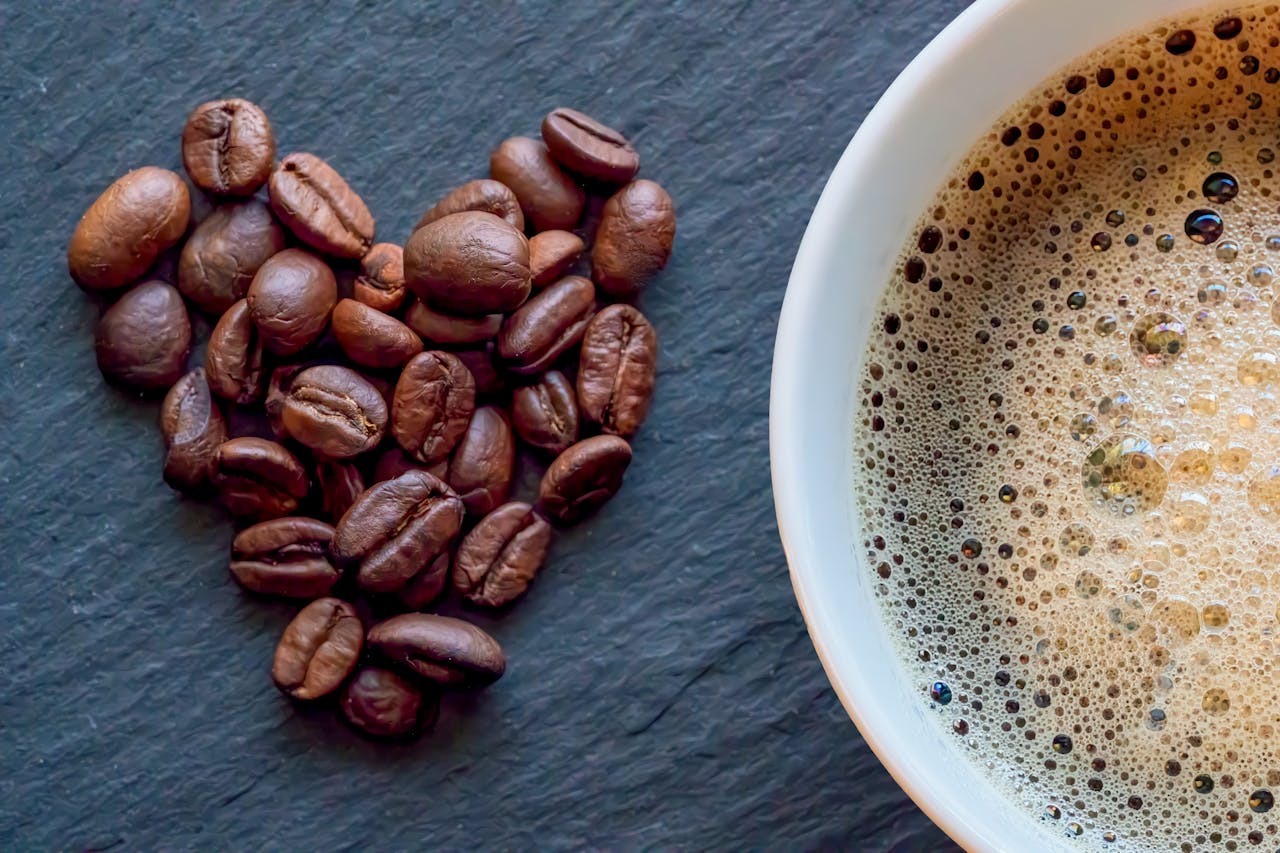services-02 Close-up of heart-shaped coffee beans next to a cup of frothy coffee on a slate surface.